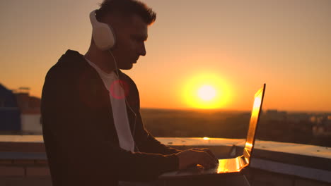 Rear-view-of-a-man-in-headphones-listening-to-music-and-working-on-the-roof-of-a-building-at-sunset-with-a-view-of-the-city-from-a-height.-Roof-of-a-skyscraper-at-sunset.